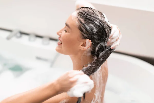 Haircare. Side View Of Woman Washing Head Caring For Long Brown Hair Sitting In Bathtub Indoors. Happy Lady Applying Shampoo Posing With Eyes Closed In Modern Bathroom, Selective Focus