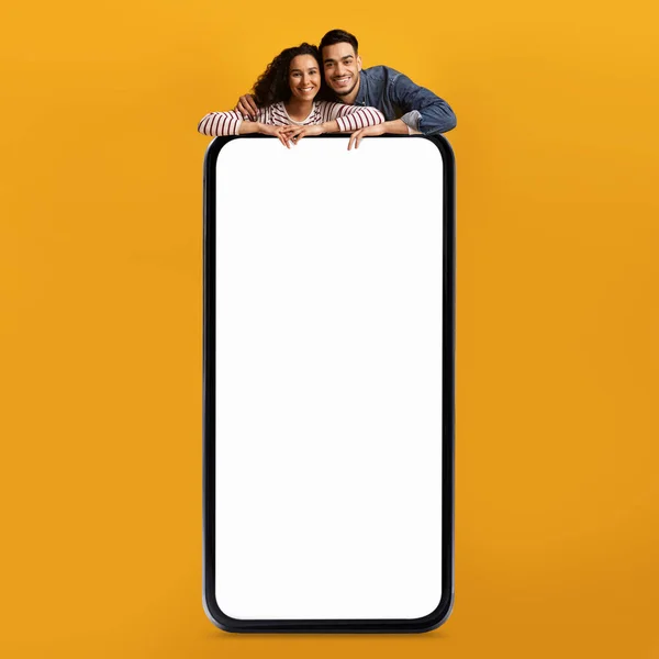 モバイル広告 黄色の背景に白い画面を持つ大きな空白のスマートフォンでリースハッピー中東のカップル オンラインオファーやアプリのデザインのためのコピースペースを実証アラブの男と女 モックアップ — ストック写真