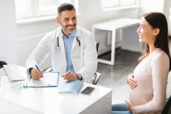 产前护理概念 快乐的年轻孕妇坐在诊所 与男妇科医生商量 女人抱着肚子 与产科医生交谈 — 图库照片