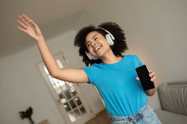 Kablosuz kulaklıklı mutlu, genç, kıvırcık, siyah kadın müzik dinler, boş zamanların tadını çıkarır, dans eder, eğlenir, oturma odasının içinde boş ekranlı bir telefon şovu yapar. Evde ses uygulaması ile eğlence