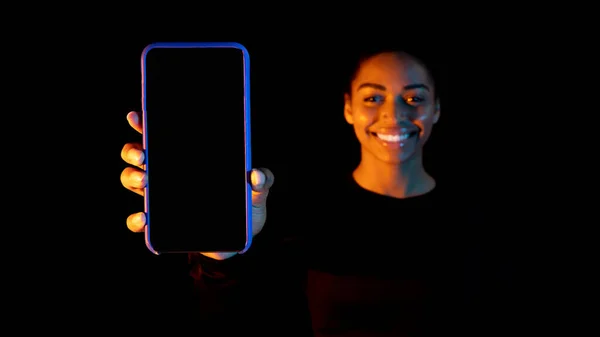 African american woman holding smartphone with black screen, posing in neon background, panorama, mockup. Happy black lady demonstrating copy space for your mobile app or website design