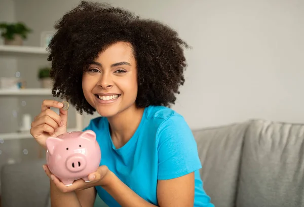 アフリカ系アメリカ人の若い女性が笑顔でコインをリビングルームのインテリアに貯金箱に入れ 自由な空間を作りました 自宅での推奨貯蓄 投資および寄付 — ストック写真