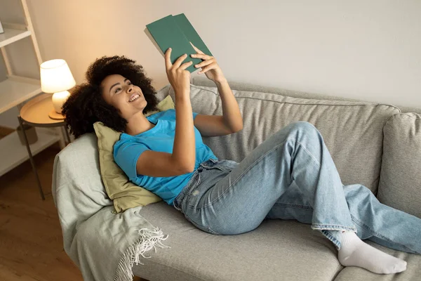 Sonriente Joven Afroamericana Rizada Mujer Leyendo Libro Encuentra Sofá Disfruta —  Fotos de Stock