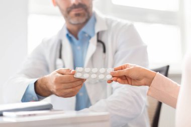 Orta yaşlı erkek doktor hamile bayana reçeteli ilaç veriyor klinikteki masada oturuyor ve tabletleri tutan ellere odaklanıyor.