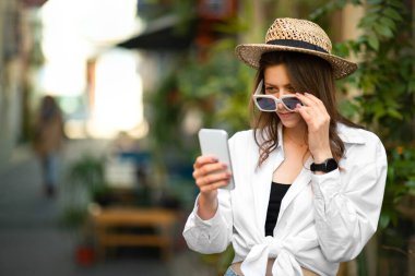 Gündelik giyinmiş, şapkalı, güneş gözlüklü, akıllı telefon haritasına bakıp yeni şehirde, dışarıda manzara arayan beyaz bir kadın. Tatil ve yaz seyahatleri, otelle ilgili sorunlar.