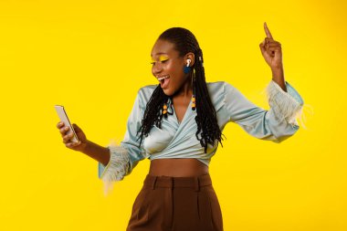 Neşeli Siyah Kadın Telefonu Tutuyor ve İnternet 'te Müzik Dinliyor Kablosuz Kulaklık Giyiyor Sarı Arkaplanda Favori Şarkı Söylüyor. Müzikal Uygulama Yönlendiricisi. Stüdyo Çekimi