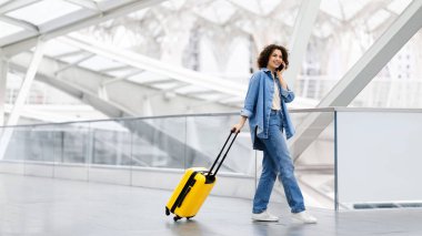 Mobil İletişim. Havaalanında cep telefonuyla konuşan mutlu genç kadın, Terminal Salonunda bavulla yürüyen güzel gülümseyen bayan ve keyifli telefon konuşmaları, kopyalama alanı.