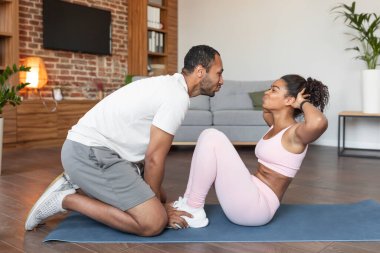 Genç ve mutlu bir zenci koca spor kıyafetlerinde karısına yardım ediyor karın kası egzersizleri yapıyor oturma odasının içinde yerde öpüşüyor. Aşk, ilişki, vücut bakımı ve destek, evde kilo kaybı ve spor