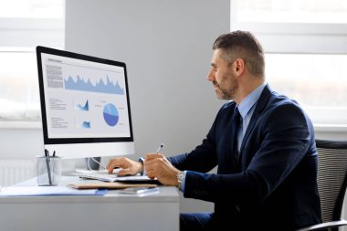 Orta yaşlı adam finans analisti istatistiksel bilgiler üzerinde düşünüyor grafiklerdeki verileri karşılaştırıyor, pazarlama stratejisine erişiyor, işyerinde oturuyor
