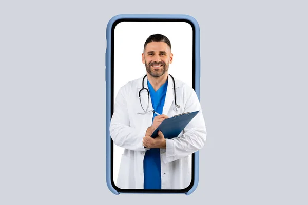 Stock image Medical services. Friendly male therapist in white uniform with stethoscope and clipboard, writing prescription to patient and smiling, looking coming out huge cellphone screen