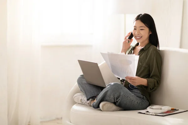 彼女の膝の上にPcコンピュータとソファの上に座っているカジュアルな服装ビジネスコンサルタントでかなり若いアジアの女性 自宅から働いて 電話を持っています 彼女の手の中に紙を見て スペースをコピー — ストック写真