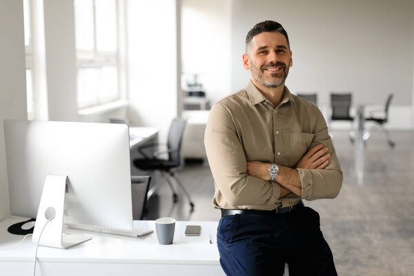 Успешный предприниматель среднего возраста, сидящий со скрещенными руками за столом в офисе, смотрящий и улыбающийся в камеру, свободное пространство. Уверенный мужчина графический дизайнер, работающий в компании