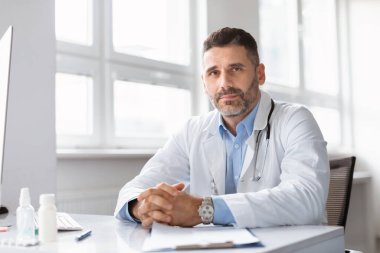 Kendine güvenen orta yaşlı erkek doktor, doktor ya da kardiyolog beyaz önlüklü adam modern ofisinde masasında oturuyor, kameraya bakıyor, boş alan arıyor.
