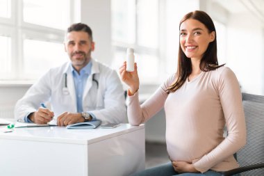 Elinde bir kavanoz hap tutan mutlu hamile kadın, kadın doğum uzmanı erkek doktorla danışma sırasında klinikte oturuyor. Doktor bayan vitaminler bekliyor.