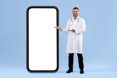 Orta yaşlı erkek doktor büyük boş bir cep telefonu ekranı gösteriyor. Mavi stüdyo arka planında poz veriyor. Boyu uzun, maketi var. Cep telefonunun yanında duran adam doktor. Tasarım şablonu