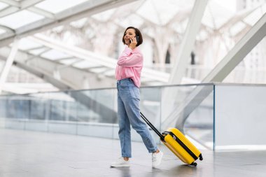 Havaalanında çantasıyla yürürken cep telefonuyla konuşan gülümseyen genç kadın, uçuş kapısına giderken cep telefonu konuşmasının keyfini çıkaran güzel milenyum kadını,