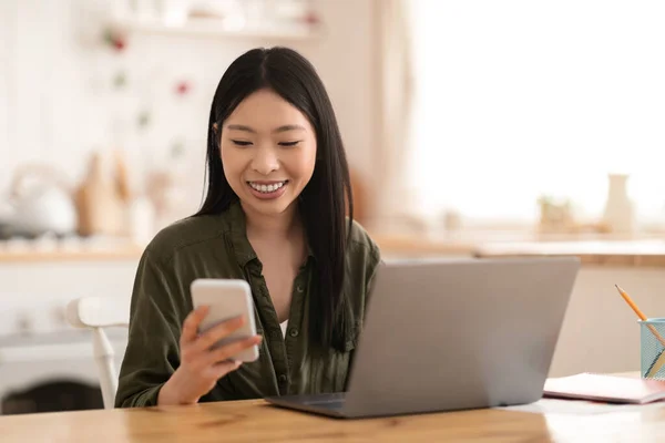 自宅で働く正の陽気なかなり若いアジアの女性のフリーランス キッチンデスクに座っている女性の独立した請負業者 ラップトップを使用してスマートフォンでメッセージをチェックし スペースをコピーします — ストック写真