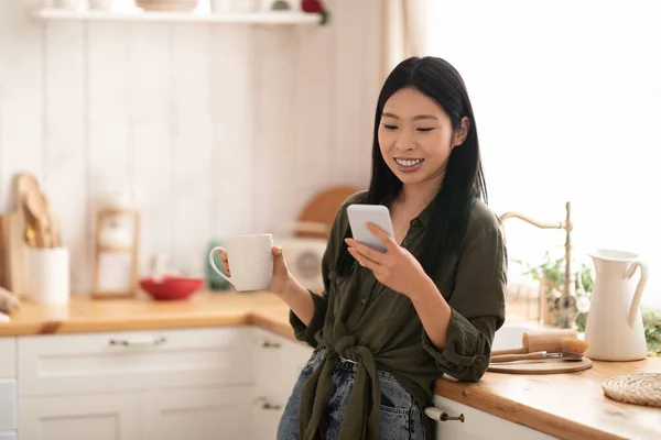 在舒适的厨房里 穿着休闲装的快乐的年轻的韩国女人站在桌子旁边 用手机笑着 拿着杯子 边喝咖啡边看消息 复制空间 — 图库照片