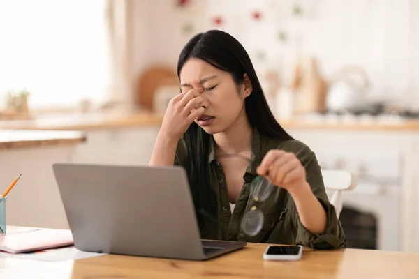 Overworked exhausted asian woman independent contractor sitting at kitchen desk in front of laptop at home, rubbing nose brisge, removing eyeglasses, feeling tired, copy space. Burnout in millennials