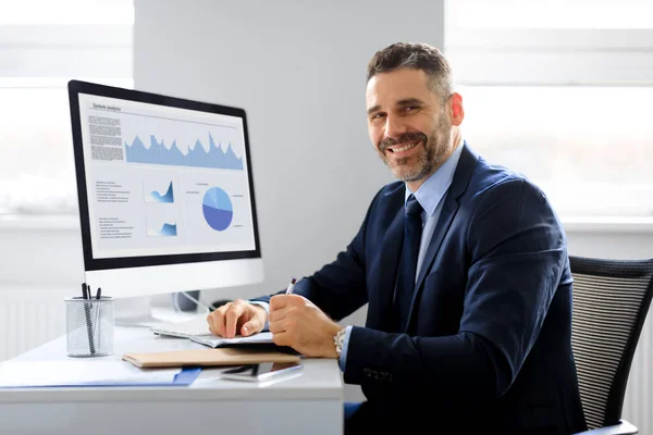 Portrait of happy businessman working on computer in office and smiling at camera, monitor with graphics and charts. Male entrepreneur working with graphs