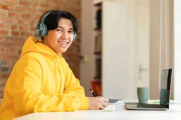 遠隔教育 中国のティーンエイジャーの男は ラップトップを使用して宿題をオンラインで行い 自宅でテーブルに座ってカメラに笑顔ノートを取ります イヤフォンを身に着けています Learning Concept — ストック写真