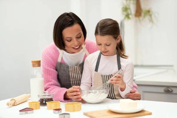 Smiling caucasian young mother and little daughter in aprons make dough for cookies with whisk in kitchen interior on table with milk and eggs. Lesson of cook food, homemade sweets at home