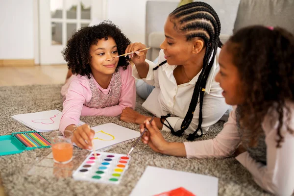 stock image Black Family Drawing And Having Fun At Home, Mom Doing Face Painting For Her Preteen Daughters, Touching Kids Nose With Brush, Bonding Spending Time Lying On Floor Indoors. Selective Focus