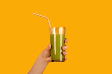 Yeşil smoothie ile el ele tutuşan tanınmayan bir kadın, turuncu stüdyo arka planında izole edilmiş, kırpılmış çekim, kopyalama alanı. Detoks, sağlıklı beslenme, enerji içeceği, vitamin ve yiyecek mineralleri.