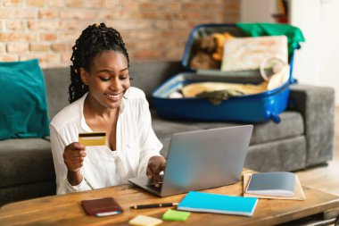 Mutlu siyahi kadın dizüstü bilgisayarını kullanıyor ve kredi kartıyla turneye çıkıyor, bavulları boşaltılmış bir şekilde koltukta oturuyor, tatile hazırlanıyor, bedava yer açıyor.