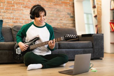 Gülümseyen Asyalı öğrenci evde yerde dizüstü bilgisayarın yanında oturup elektro gitarla müzik çalıyor. Çocuk, hafta sonları İnternet üzerinden akorları öğreniyor. Hobi ve Hobi Konsepti. Seçici Odaklanma