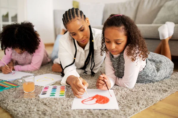 Black Mom And Two Preteen Daughters Painting Heart Spending Time Together Lying On Floor At Home. Family Bonding Sketching And Drawing Together On Weekend. Leisure Activity And Hobby