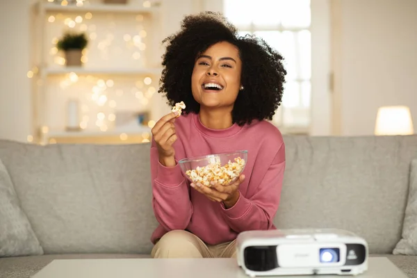 Happy Black Woman Using Movie Projector Watching Film And Eating Popcorn Having Fun Sitting On Couch At Home. Weekend Leisure And Domestic Gadgets Concept. Selective Focus