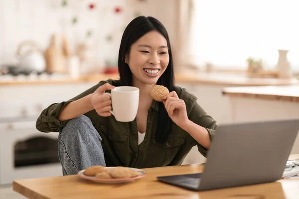 Joyful happy pretty young asian woman in casual relaxing in kitchen, sitting at desk in front of laptop, drinking coffee, eating cookies while watching movie or video content on Internet, copy space