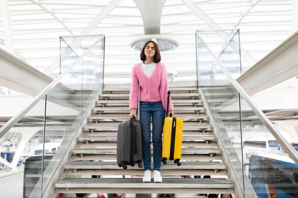 Upset young woman walking down stairs at airport with heavy suitcases, millennial female travelling alone, carrying luggage and frowning while going to flight departure gate in terminal, copy space