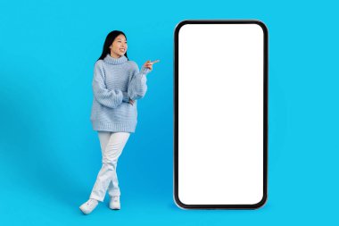 Hoş, uzun saçlı, sıcak süveter ve beyaz pantolonlu, mavi stüdyo arka planında poz veren, güzel bir teklif sunan, boş ekran modeliyle büyük bir telefona bakan, uzun boylu Asyalı bir kadın.