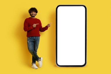Havalı, yakışıklı, gündelik kıyafetli, beyaz ekranlı, büyük cep telefonunu işaret eden, sarı stüdyo arka planında izole edilmiş, taklit alanı, taklit alanı olan, güzel bir teklif sunan yakışıklı doğulu adam.