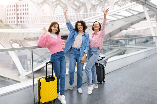 空港でポーズをとりながらカメラで手を振っている3人の幸せな女友達のポートレート 陽気な若い女性が一緒に旅行し 出発を待っている間にターミナルでスーツケースと一緒に立ちます — ストック写真