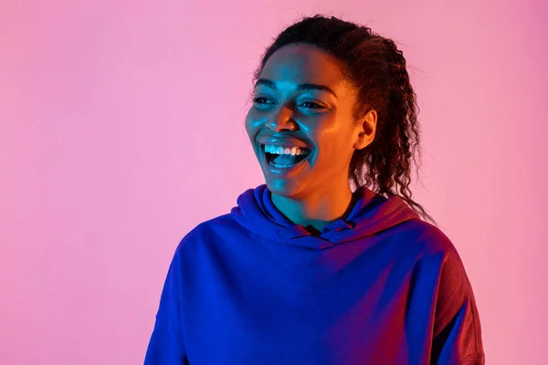 Portrait of happy young african american woman looking aside and smiling in pink neon light background, copy space. Attractive black lady expressing positivity, feeling cheerful