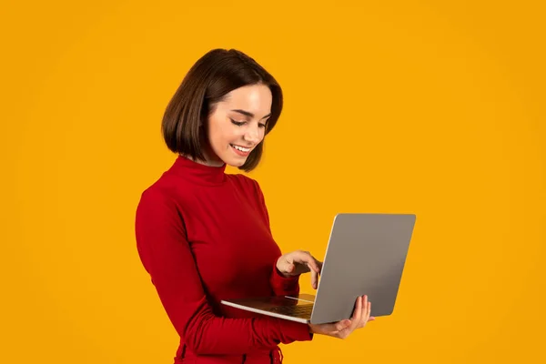 远程工作 自由职业概念 穿着红衣服 面带微笑的漂亮女人 在笔记本电脑键盘上用漂亮的发型打字 远程工作 在橙色工作室背景下与世隔绝 — 图库照片