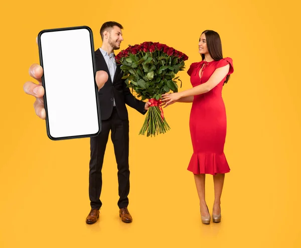 浪漫的男人向女朋友送花 在相机前展示大大的空白智能手机 推荐网上购物应用程序 并送上情人节礼物 站在黄色工作室的背景上 — 图库照片