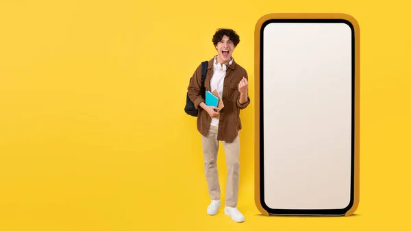情绪激动的男生站在智能手机旁边 用巨大的空屏幕 在黄色工作室的背景下高喊和显示 是的广告教育移动应用 — 图库照片