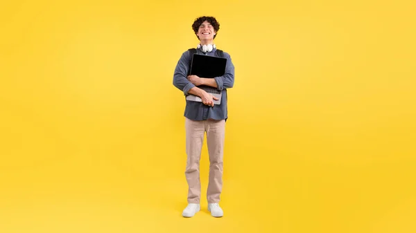 电子学习 快乐的男生抱着他的新笔记本电脑 背着背包站在黄色工作室的背景下 紧闭双眼的家伙紧紧抓住电脑广告教育网站 — 图库照片