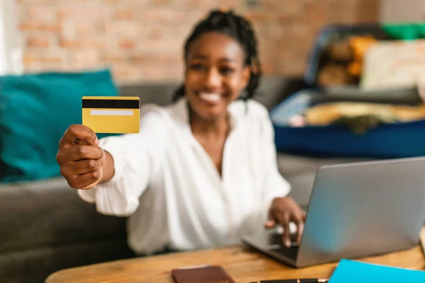Happy black woman using laptop and booking hotel online with credit card, selective focus on bank card, closeup. Lady sitting with suitcase, ready to go