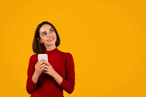 stock image Curious happy pretty young brunette woman in red using smartphone on orange studio background, looking at copy space and smiling, checking exciting online offer or nice ad