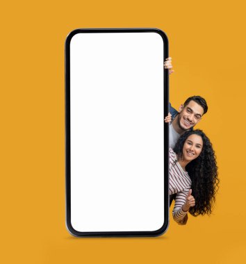 Mobil Model. Neşeli bir Arap çift, büyük, boş, akıllı bir telefonun arkasından bakıp sarı arka planda başparmağını kaldırıyor, mutlu bir Orta Doğulu erkek ve kadın, yeni bir uygulama ya da web sitesi öneriyor,