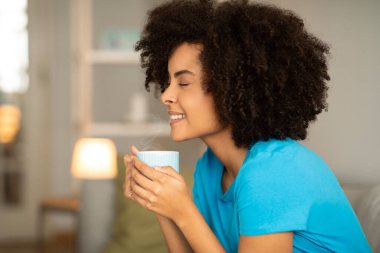 Bir fincan sıcak içecekle mutlu, kıvırcık, Afro-Amerikalı bir kadın. Oturma odasının içinde rahatlama ve aromanın tadını çıkar. Kahve molası, evde çay molası.