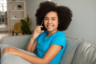 Gülümseyen kıvırcık, Afro-Amerikan kadın akıllı telefondan arıyor, gri oturma odasının içinde konuşmaktan hoşlanıyor. Cihazla iletişim, dedikodu ve iyi haberler, evde yeni normaller