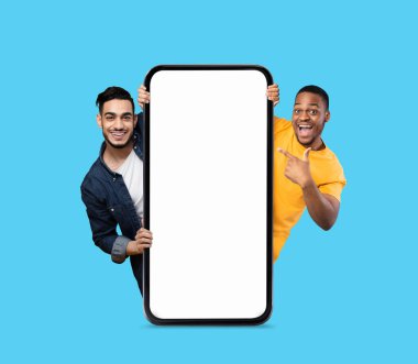 Mavi arkaplanı beyaz ekranlı, beyaz akıllı telefonu işaret eden iki neşeli adam, mutlu Arap erkek ve siyahi adam online teklif veya mobil uygulama, kolaj,