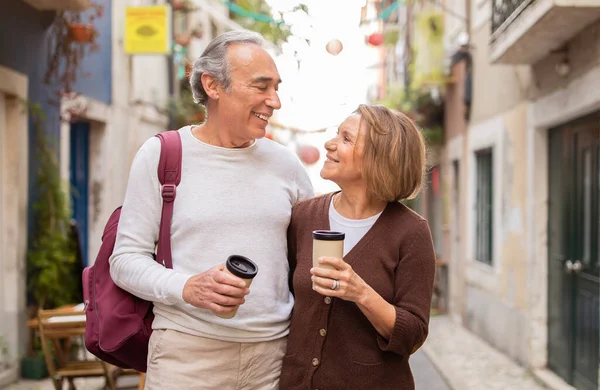 Retirement Travel. Cheerful Senior Couple Walking With Backpack Drinking Coffee In Paper Cups Enjoying Sightseeing And Vacation In European City Outside. Happy Retired Lifestyle Concept