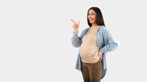 いい提案だ 無料のスペースで脇を指して カメラで笑顔 妊娠中の女性は白いスタジオの背景にデザインのための無料の場所を示す コピースペースとパノラマ — ストック写真